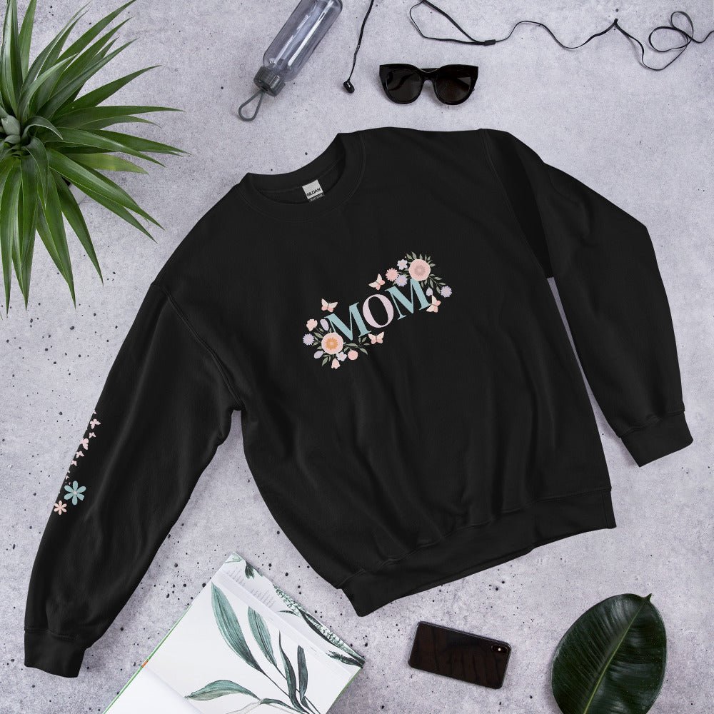 Black sweatshirt with pastel floral “MOM” lettering, laid flat on a cozy lifestyle scene with sunglasses, plants, phone, and journal – stylish gift idea for moms.