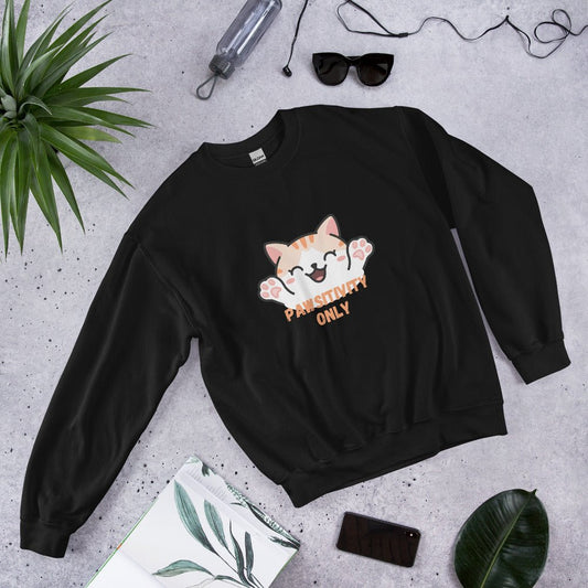 Black crewneck sweatshirt with a cute orange cat and “Pawsitivity Only” text, laid flat on a stone surface surrounded by sunglasses, a plant, phone, and notebook.