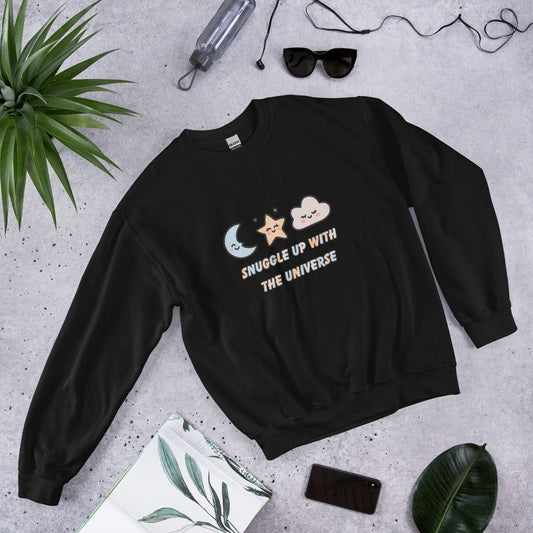 Black sweatshirt with the phrase “Snuggle Up with the Universe” and illustrations of a moon, star, and clouds, laid flat on a concrete background next to sunglasses, a water bottle, and tropical leaves.