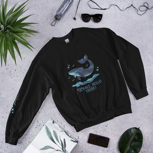 Flat lay of a black sweatshirt with a cute whale and ocean wave print, reading “Whale, Hello There!” – styled with sunglasses, a water bottle, and tropical leaves on a concrete background.