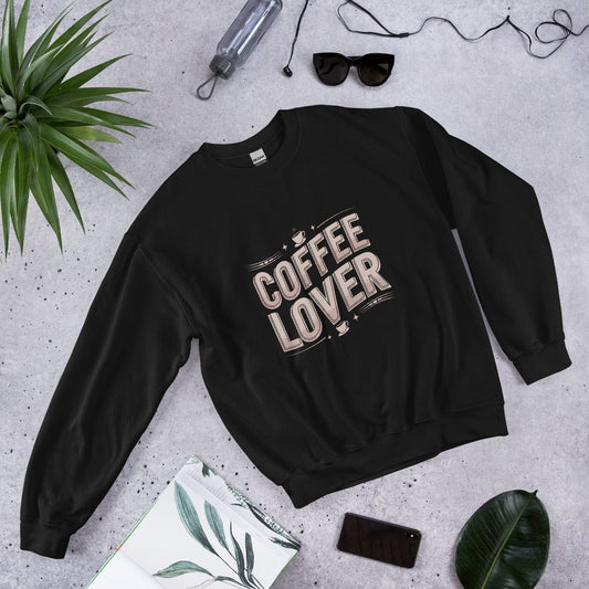 Flat lay of a black crewneck sweatshirt with bold beige lettering that reads "COFFEE LOVER," surrounded by accessories like sunglasses, a water bottle, and green plants, styled on a light gray background.