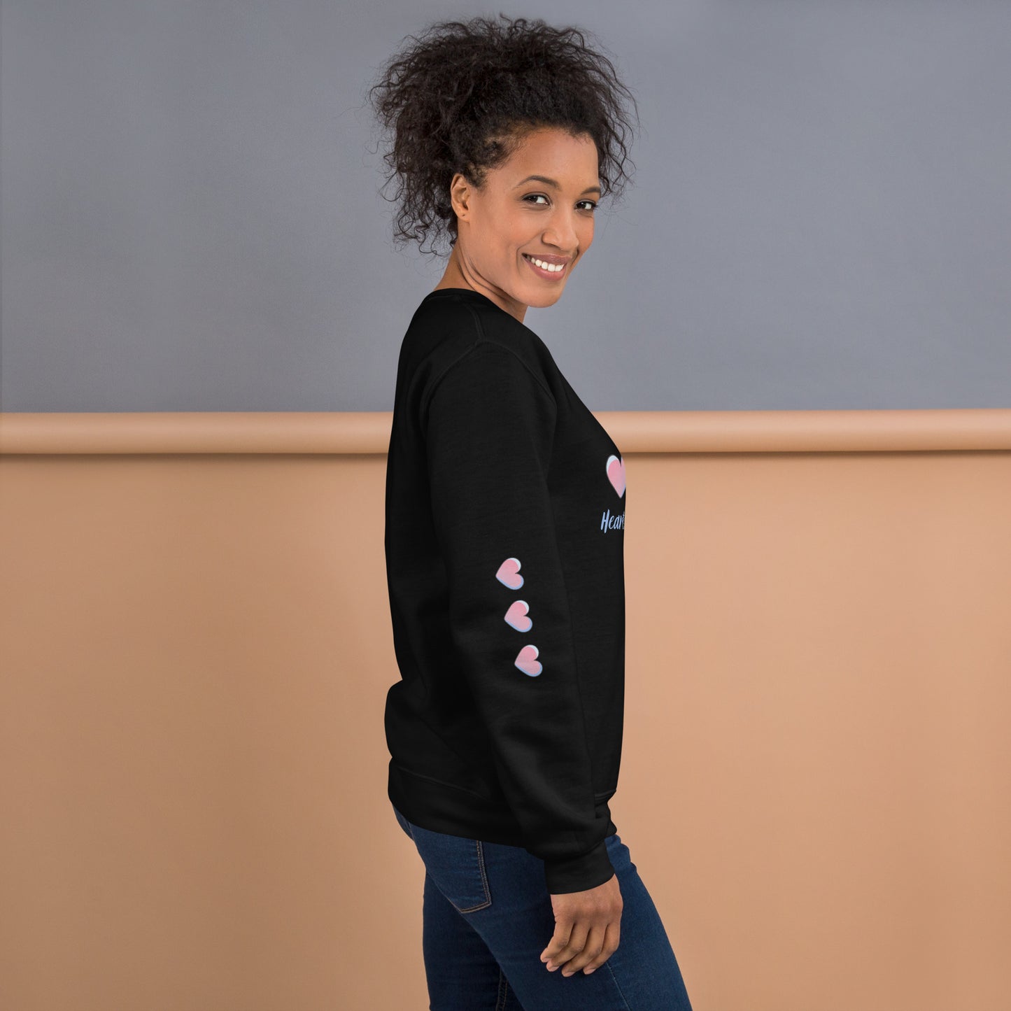 Smiling woman wearing a cozy black sweatshirt with three pink hearts running down the sleeve – side view showcasing playful and inspirational mom-style loungewear.