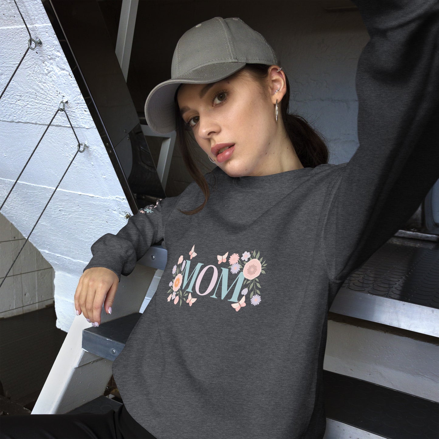 Young woman in a gray baseball cap posing in a cozy dark heather sweatshirt with pastel floral “MOM” design – casual street-style vibe with a soft, feminine twist.