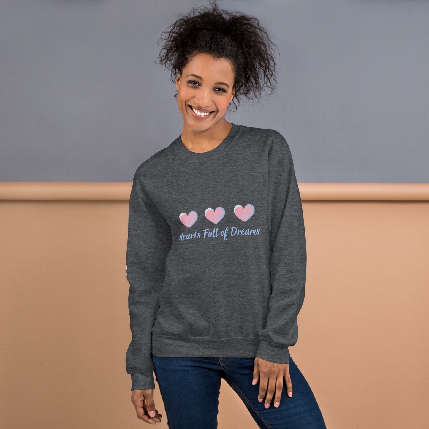Smiling woman wearing a dark heather sweatshirt with a pastel heart design and the phrase “Hearts Full of Dreams”
