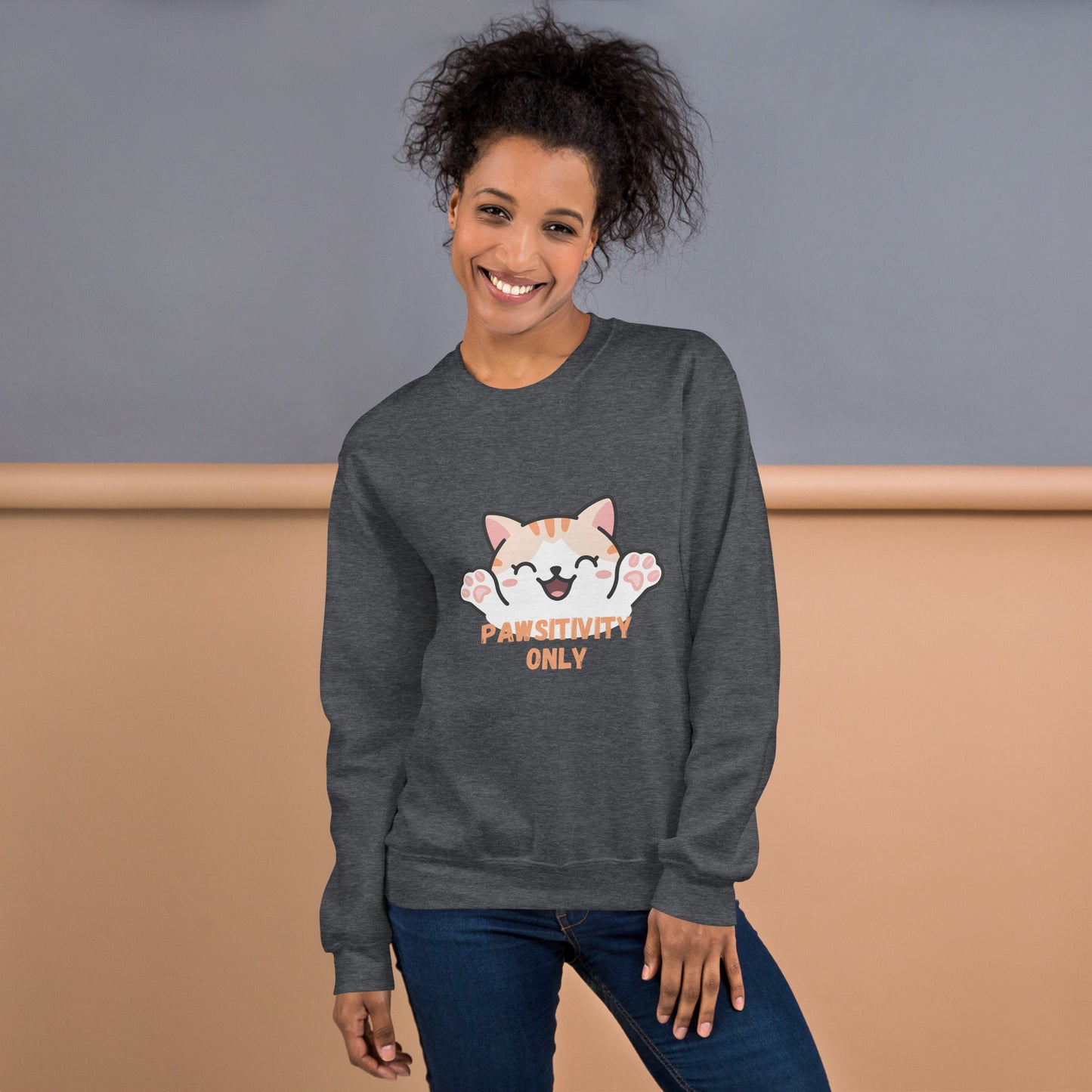 Smiling woman wearing a dark heather sweatshirt with a cute orange cartoon cat and text that says 'Pawsitivity Only' on the front.