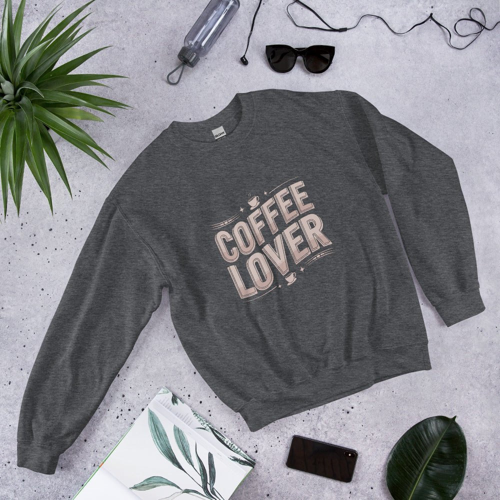 Flat lay of a dark heather crewneck sweatshirt with bold beige lettering that reads "COFFEE LOVER" surrounded by accessories like sunglasses, a water bottle, and green plants, styled on a light gray background.