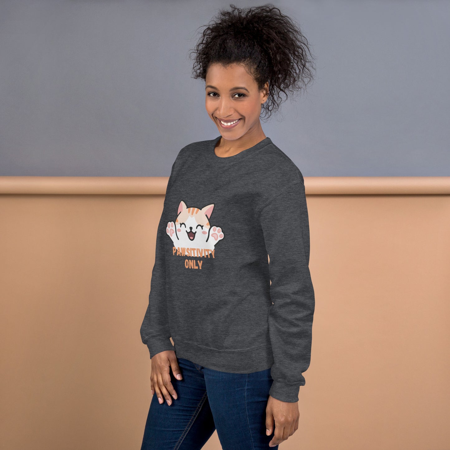 Side view of a smiling woman wearing a dark heather crewneck sweatshirt with a cute cat design and text “Pawsitivity Only” .
