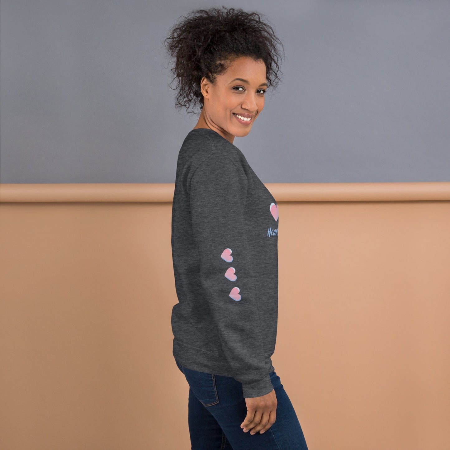Smiling woman wearing a cozy dark heahter sweatshirt with three pink hearts running down the sleeve – side view showcasing playful and inspirational mom-style loungewear.