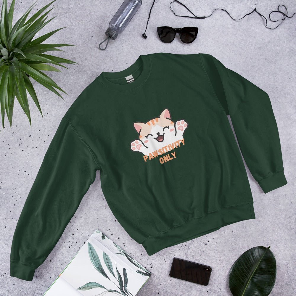 Forest green crewneck sweatshirt with a cute orange cat and “Pawsitivity Only” text, laid flat on a stone surface surrounded by sunglasses, a plant, phone, and notebook.