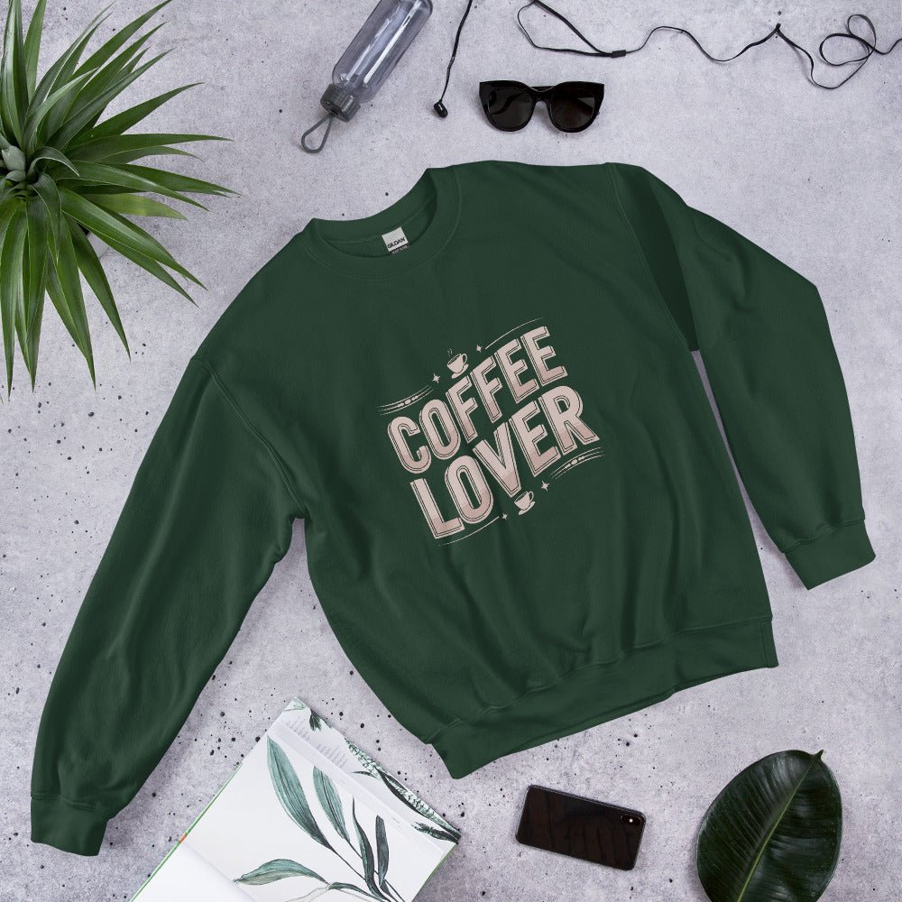 Flat lay of a forest green crewneck sweatshirt with bold beige lettering that reads "COFFEE LOVER," surrounded by accessories like sunglasses, a water bottle, and green plants, styled on a light gray background.