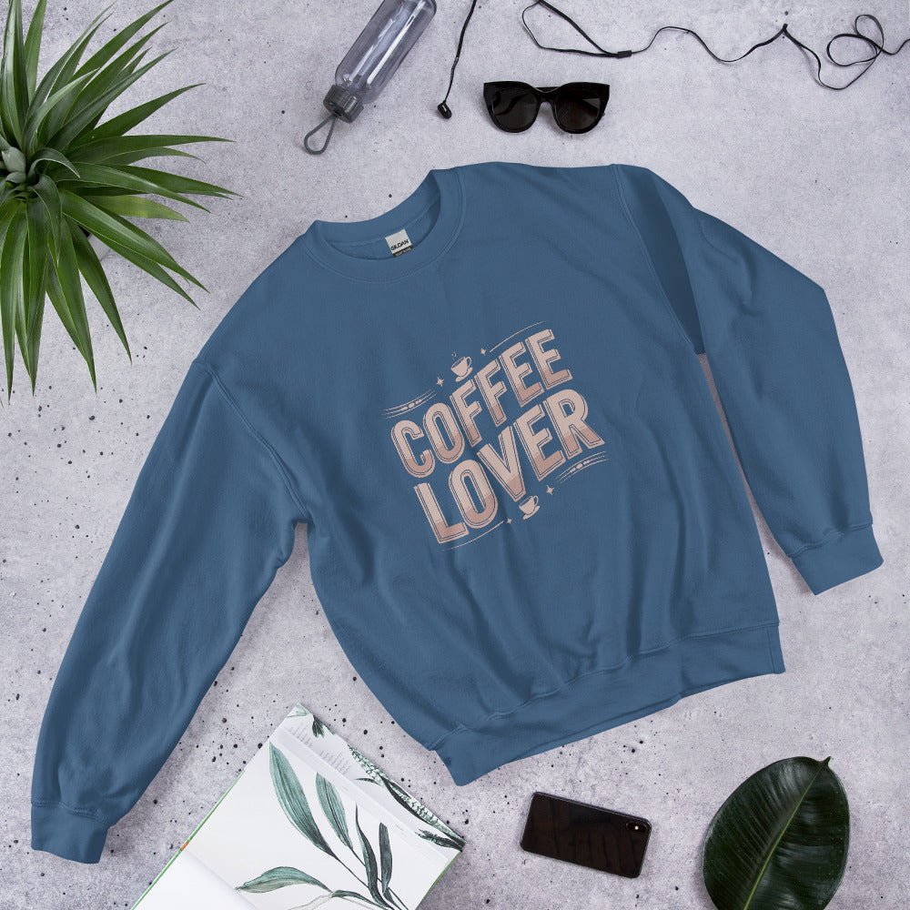Flat lay of a indigo blue crewneck sweatshirt with bold beige lettering that reads "COFFEE LOVER," surrounded by accessories like sunglasses, a water bottle, and green plants, styled on a light gray background.