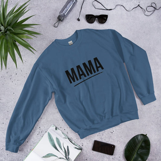 Flat lay of indigo blue MAMA sweatshirt with bold black text and underline, surrounded by sunglasses, a plant leaf, notebook, and smartphone