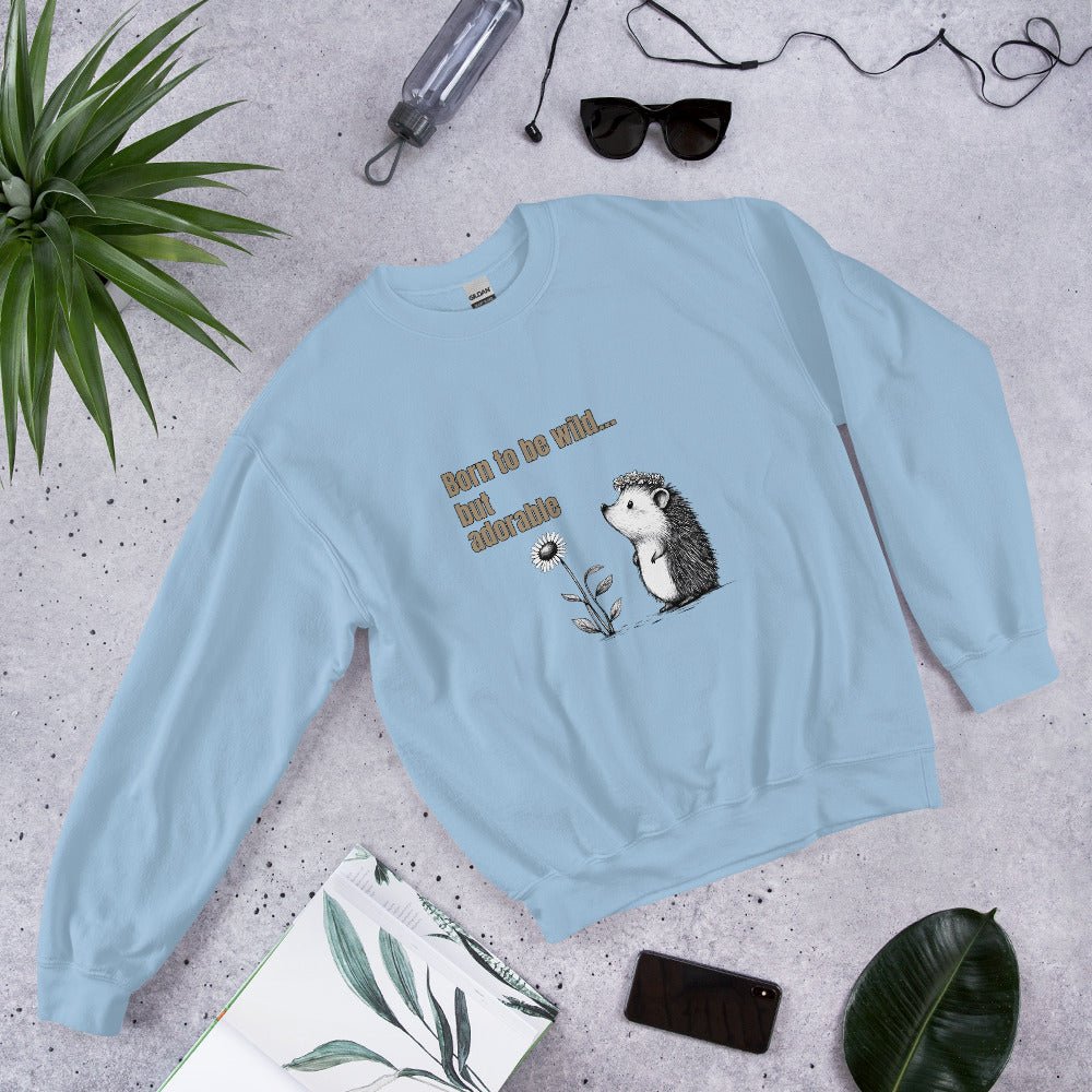 Light blue sweatshirt with a cute hedgehog and “Born to be wild… but adorable” text, styled flat lay with sunglasses, phone, and green leaves.