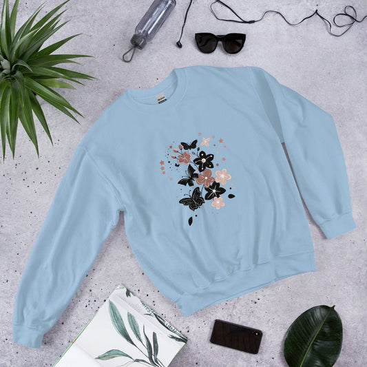 Flat lay of a light blue sweatshirt with a black and pink floral butterfly design, styled with sunglasses, a water bottle, and leaves on a light stone background.