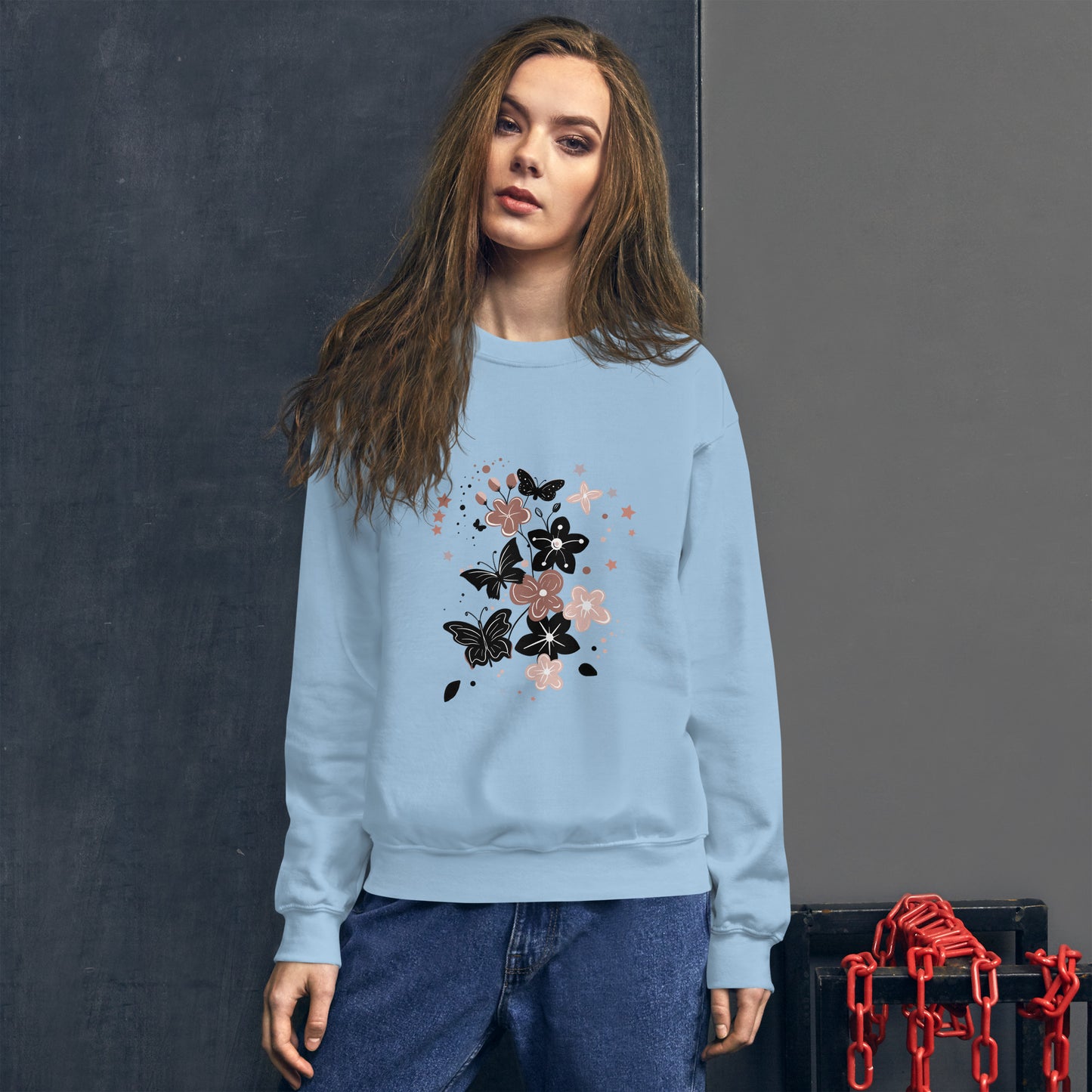 Woman wearing a light blue sweatshirt with a black and pink floral butterfly design on the chest, standing in front of a dark textured wall.