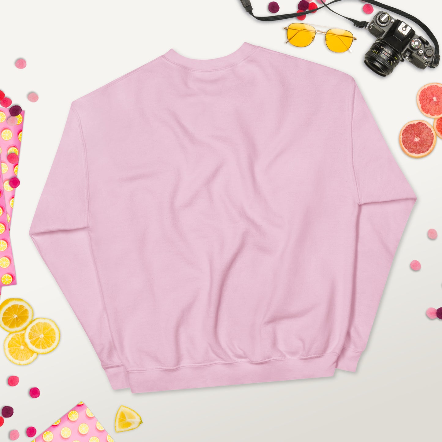 Back view of a soft pink unisex crew neck sweatshirt laid flat on a colorful background with fruits, sunglasses, and a camera.