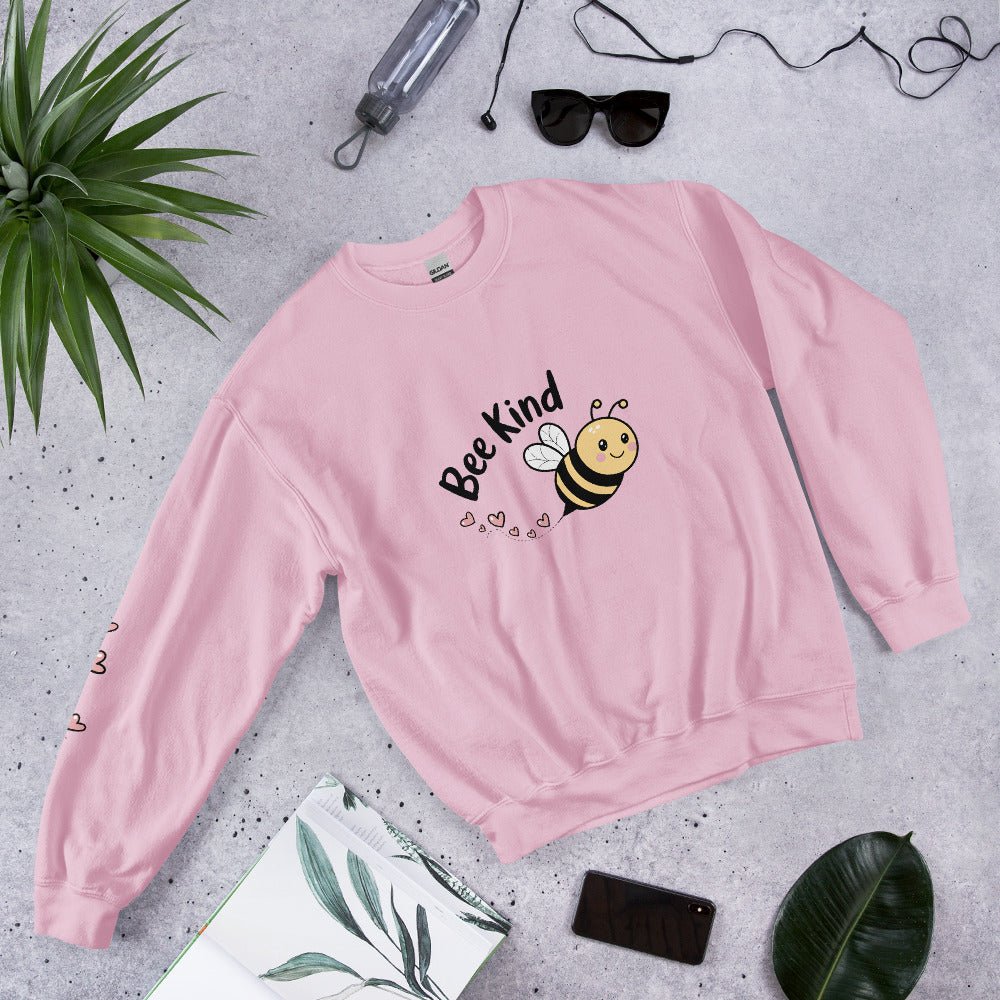 Flat lay of a cozy soft pink “Bee Kind” sweatshirt surrounded by tropical leaves, sunglasses, and a water bottle