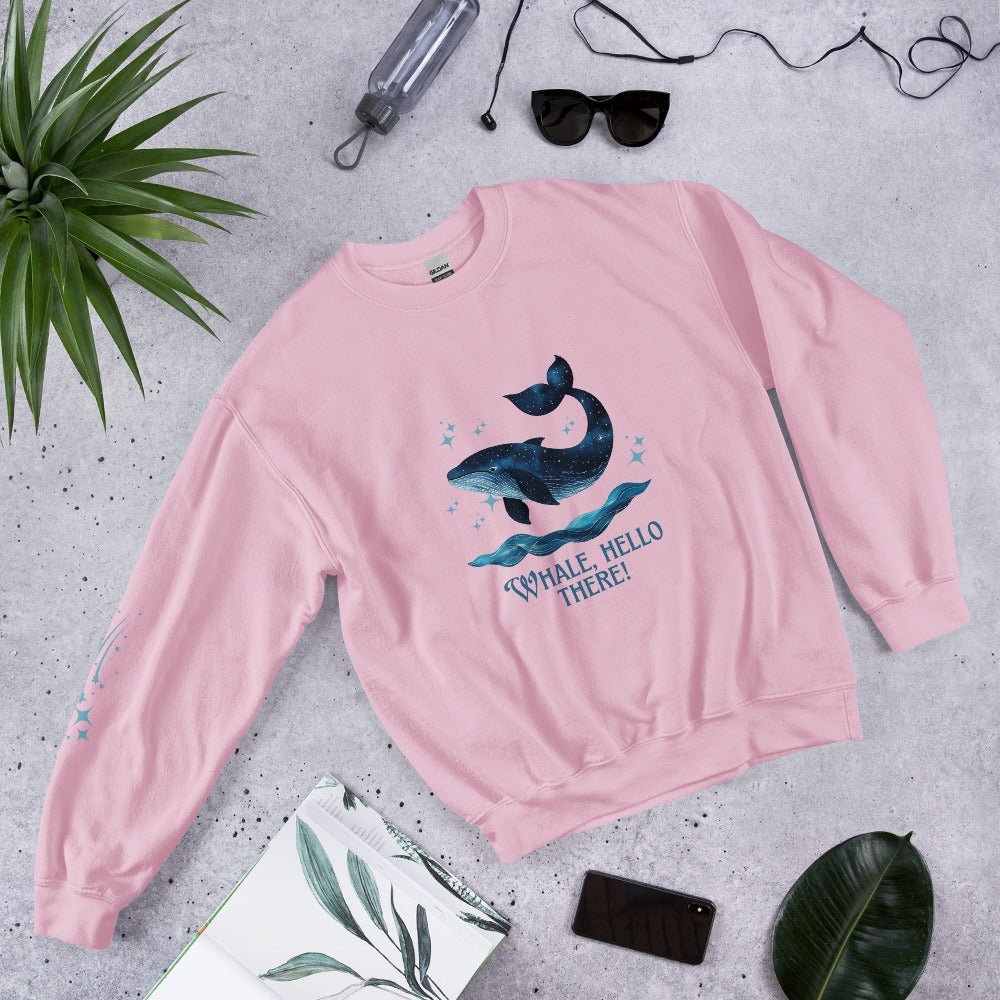 Flat lay of a soft pink sweatshirt with a cute whale and ocean wave print, reading “Whale, Hello There!” – styled with sunglasses, a water bottle, and tropical leaves on a concrete background.