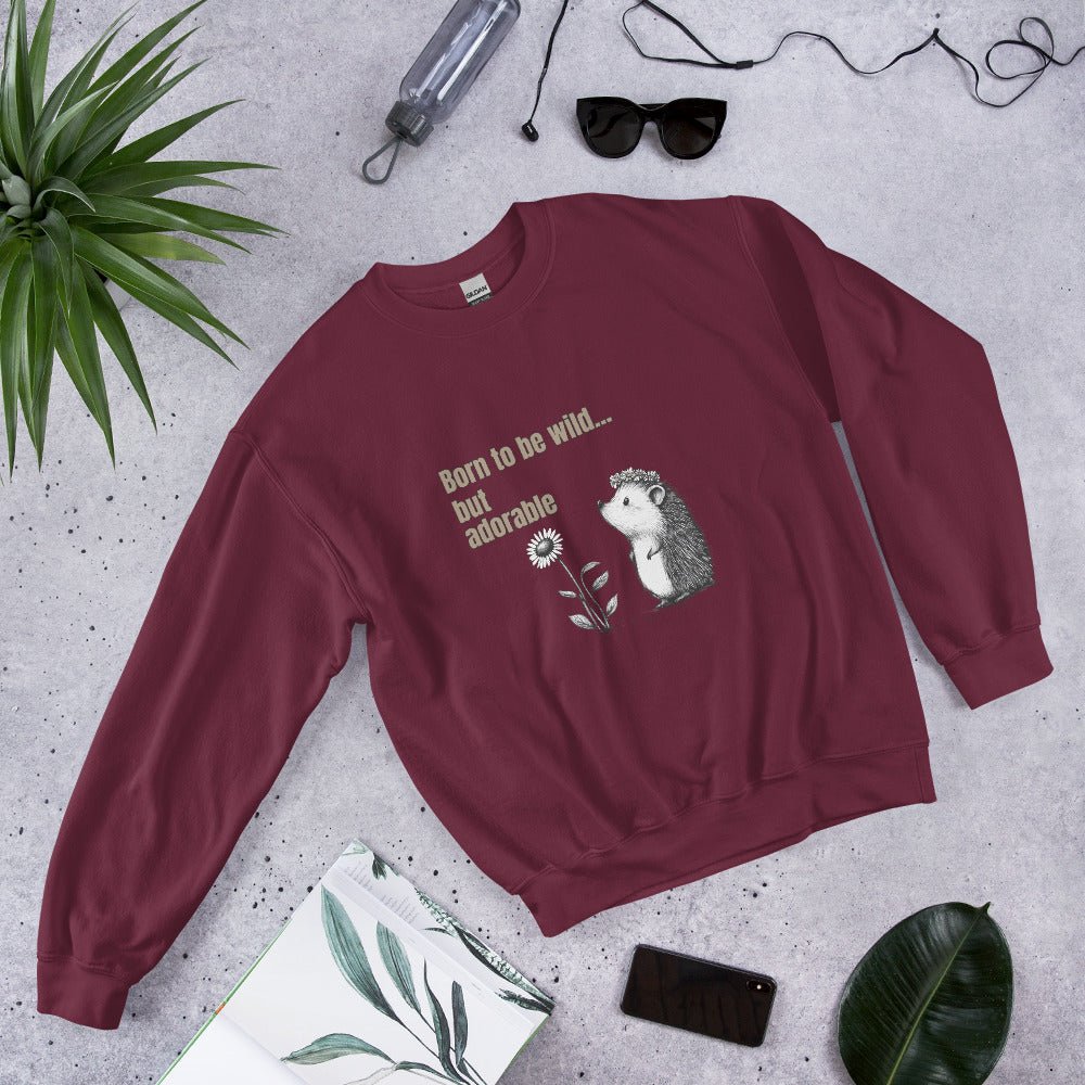 Maroon sweatshirt with a cute hedgehog and “Born to be wild… but adorable” text, styled flat lay with sunglasses, phone, and green leaves.