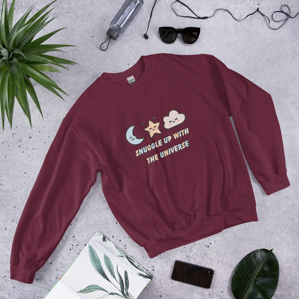Maroon red sweatshirt with the phrase “Snuggle Up with the Universe” and illustrations of a moon, star, and clouds, laid flat on a concrete background next to sunglasses, a water bottle, and tropical leaves.