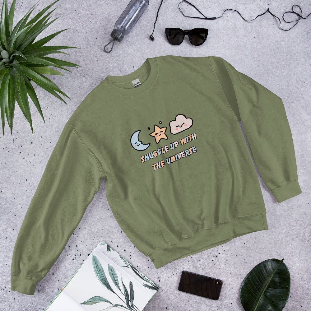 Military green sweatshirt with the phrase “Snuggle Up with the Universe” and illustrations of a moon, star, and clouds, laid flat on a concrete background next to sunglasses, a water bottle, and tropical leaves.