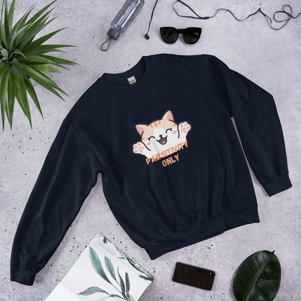 Navy blue crewneck sweatshirt with a cute orange cat and “Pawsitivity Only” text, laid flat on a stone surface surrounded by sunglasses, a plant, phone, and notebook.
