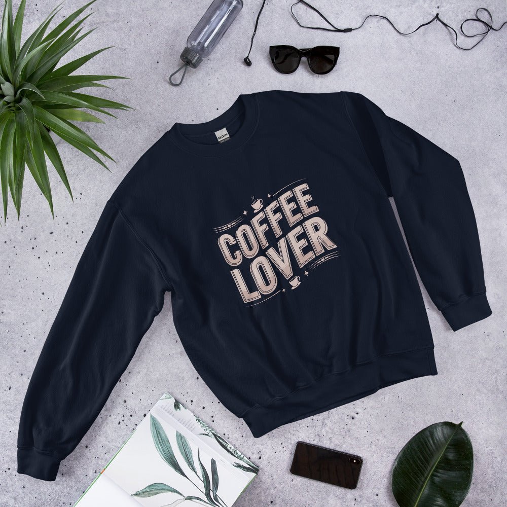 Flat lay of a navy blue crewneck sweatshirt with bold beige lettering that reads "COFFEE LOVER," surrounded by accessories like sunglasses, a water bottle, and green plants, styled on a light gray background.