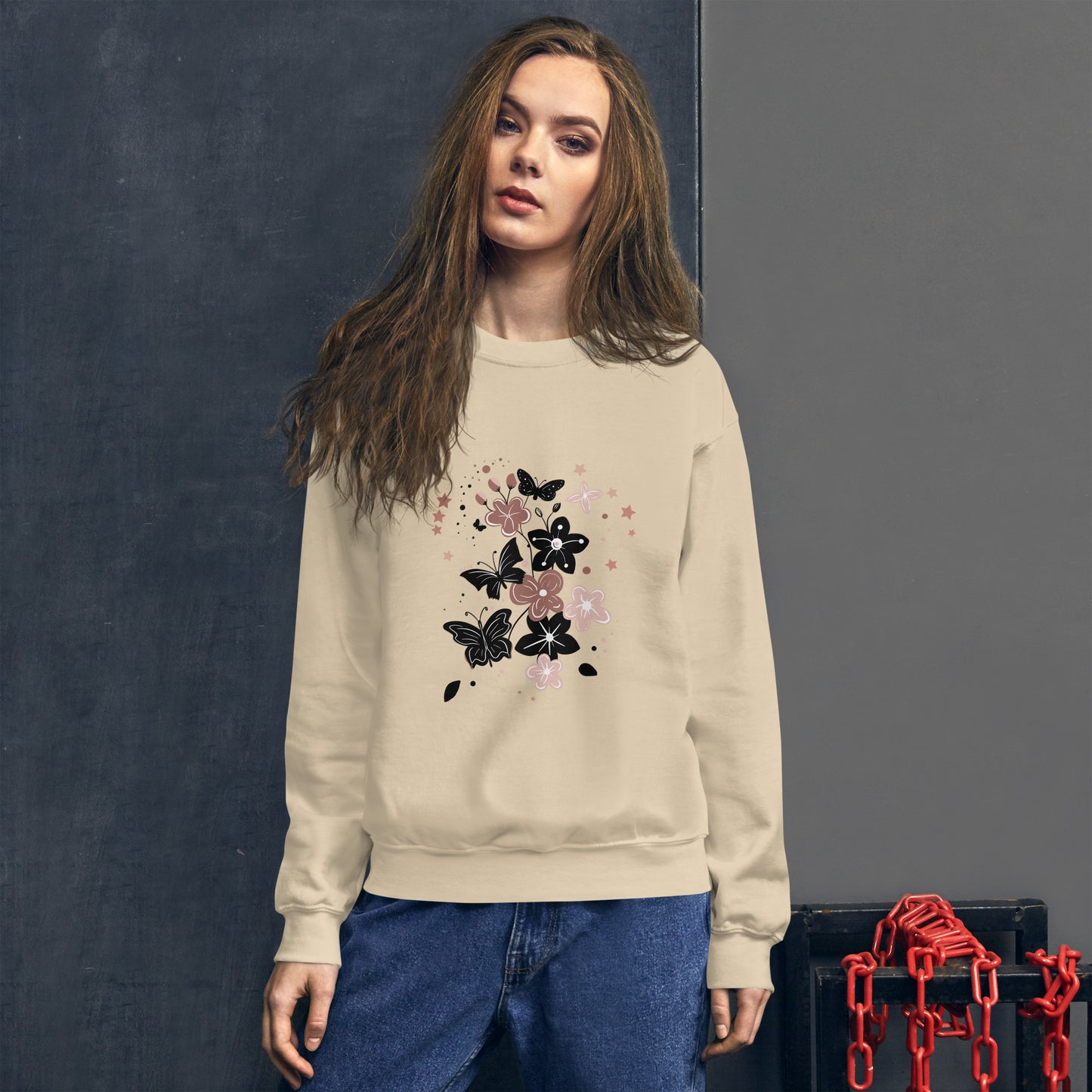 Woman wearing a sand color sweatshirt with a black and pink floral butterfly design on the chest, standing in front of a dark textured wall.