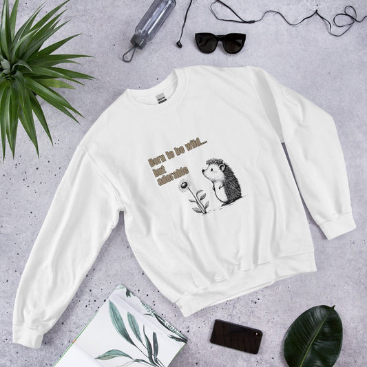 White sweatshirt with a cute hedgehog and “Born to be wild… but adorable” text, styled flat lay with sunglasses, phone, and green leaves.