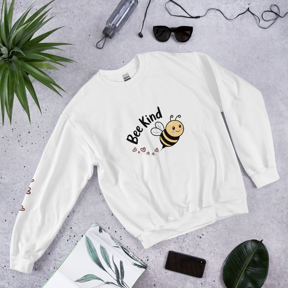 Flat lay of a cozy white “Bee Kind” sweatshirt surrounded by tropical leaves, sunglasses, and a water bottle