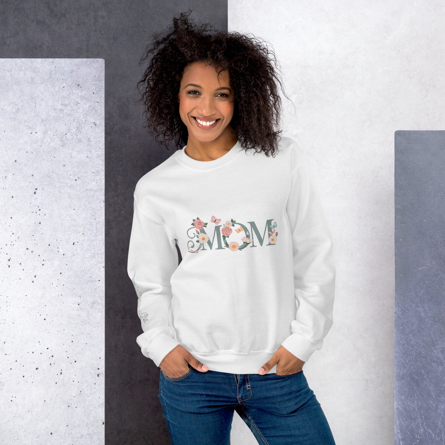 Smiling woman wearing a white floral "MOM" sweatshirt, standing against a modern textured background – cozy and stylish gift for moms.