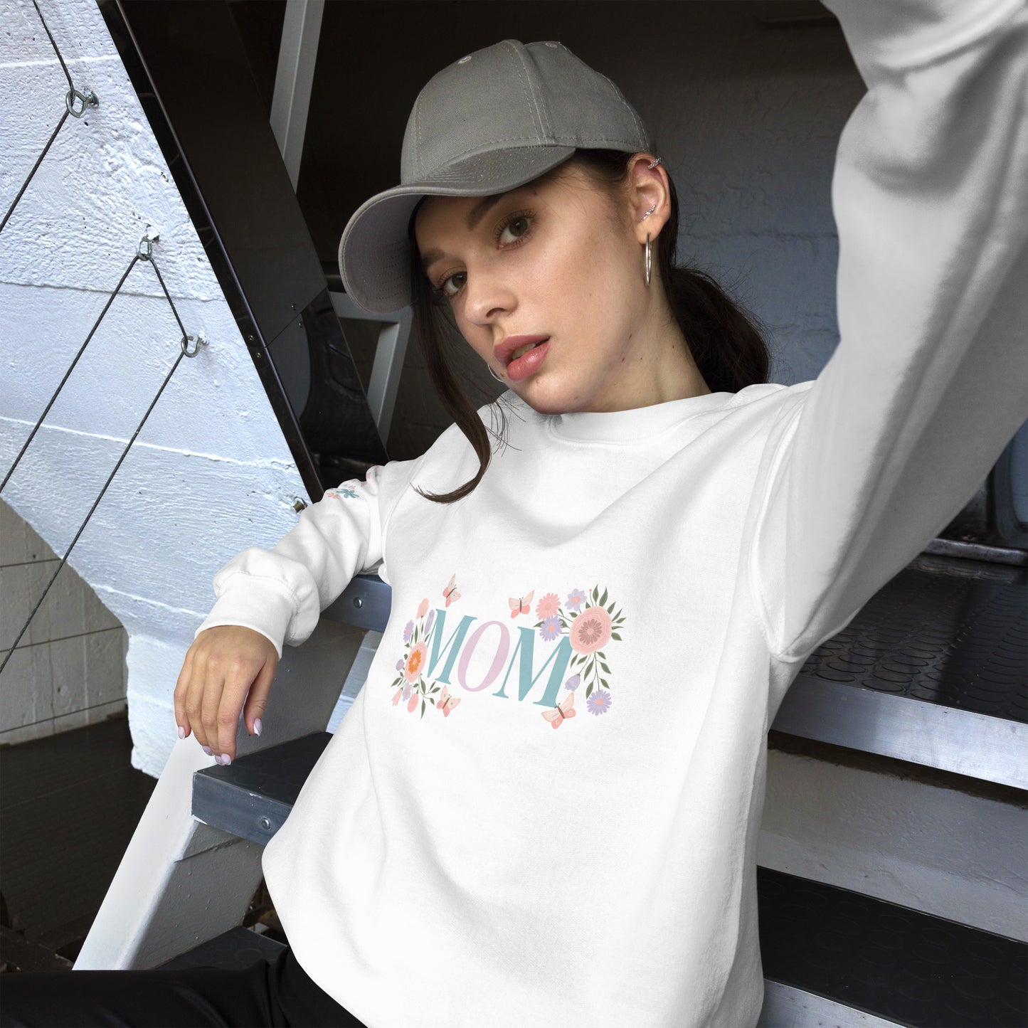Young woman in a gray baseball cap posing in a cozy white sweatshirt with pastel floral “MOM” design – casual street-style vibe with a soft, feminine twist.