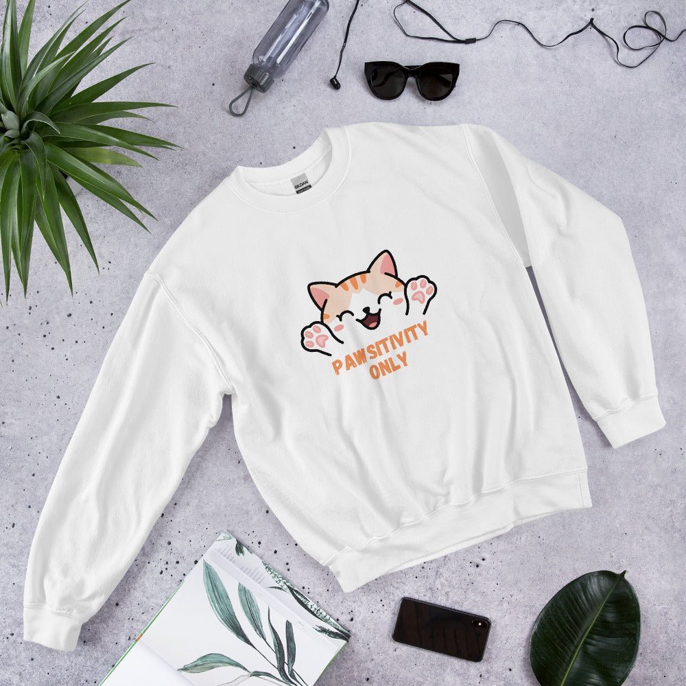 White crewneck sweatshirt with a cute orange cat and “Pawsitivity Only” text, laid flat on a stone surface surrounded by sunglasses, a plant, phone, and notebook.
