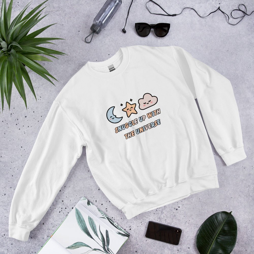 White sweatshirt with the phrase “Snuggle Up with the Universe” and illustrations of a moon, star, and clouds, laid flat on a concrete background next to sunglasses, a water bottle, and tropical leaves.