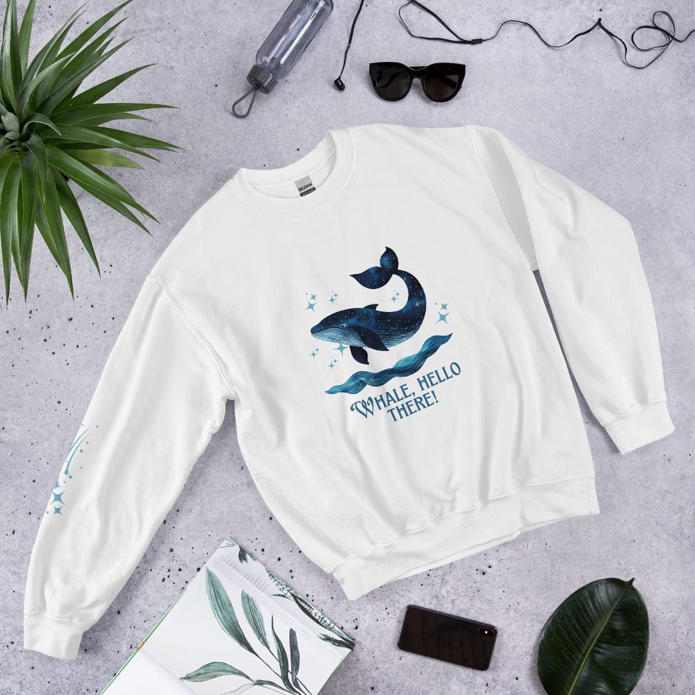 Flat lay of a white sweatshirt with a cute whale and ocean wave print, reading “Whale, Hello There!” – styled with sunglasses, a water bottle, and tropical leaves on a concrete background.