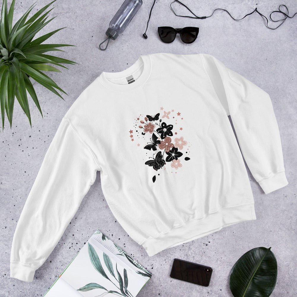 Flat lay of a white sweatshirt with a black and pink floral butterfly design, styled with sunglasses, a water bottle, and leaves on a light stone background.