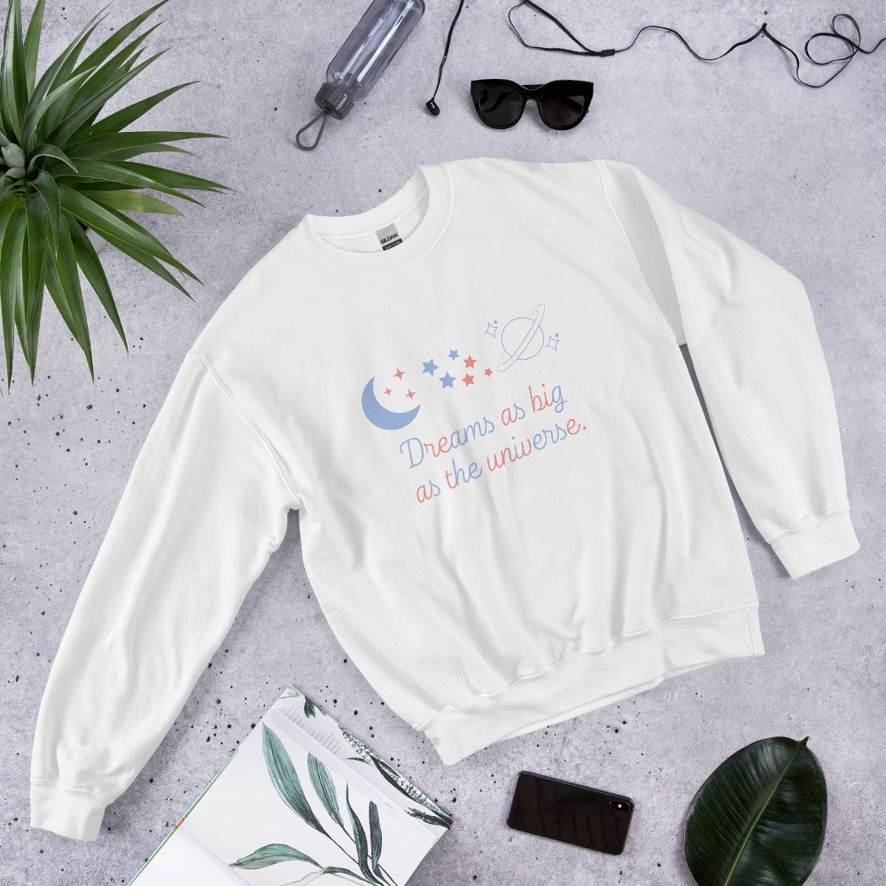 Flat lay of a white unisex Gildan sweatshirt with a pastel space-themed “Dreams as big as the universe” design, styled with sunglasses, a notebook, a phone, and tropical leaves on a gray stone background.