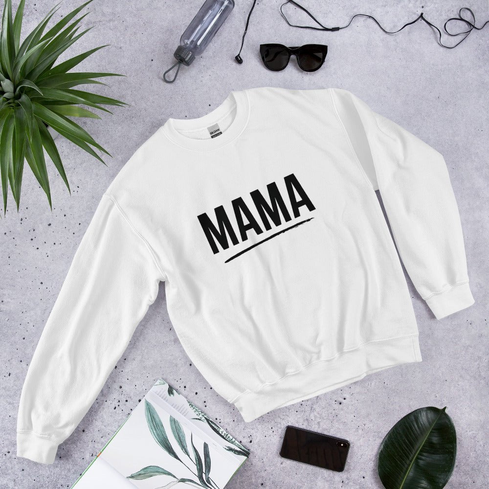 Flat lay of white MAMA sweatshirt with bold black text and underline, surrounded by sunglasses, a plant leaf, notebook, and smartphone
