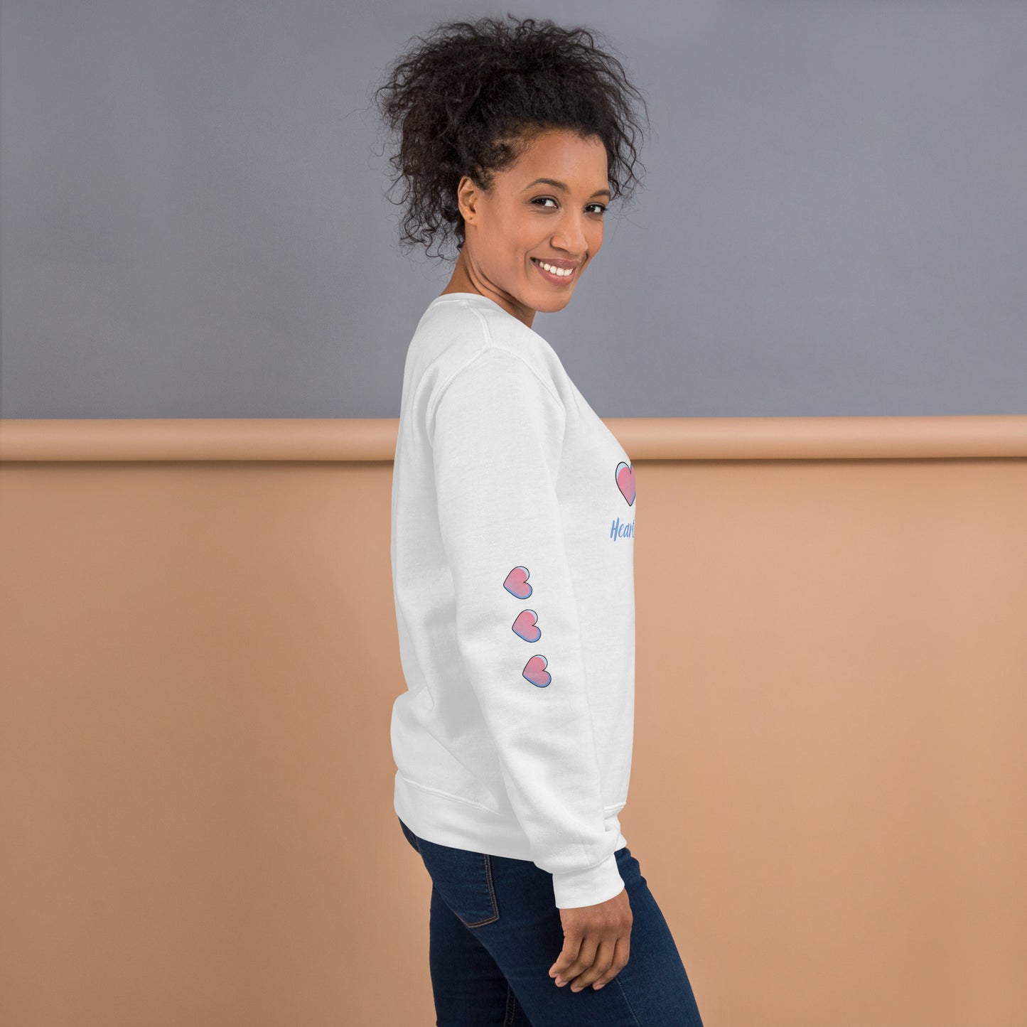Smiling woman wearing a cozy white sweatshirt with three pink hearts running down the sleeve – side view showcasing playful and inspirational mom-style loungewear.