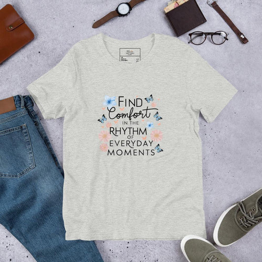 Heather gray unisex t-shirt laid flat with the quote “Find Comfort in the Rhythm of Everyday Moments,” surrounded by blue butterflies and pink flowers, styled with jeans, sneakers, a watch, glasses, and a notebook.