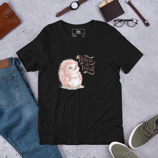 Flat lay of a black t-shirt with a pastel hedgehog and “Believe in Yourself” quote – cozy motivational tee styled with jeans and accessories.