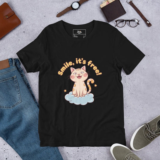 Black t-shirt laid flat on a surface, featuring a cute cat sitting on a cloud with the phrase ‘Smile, it’s free!’ surrounded by casual clothing and accessories.
