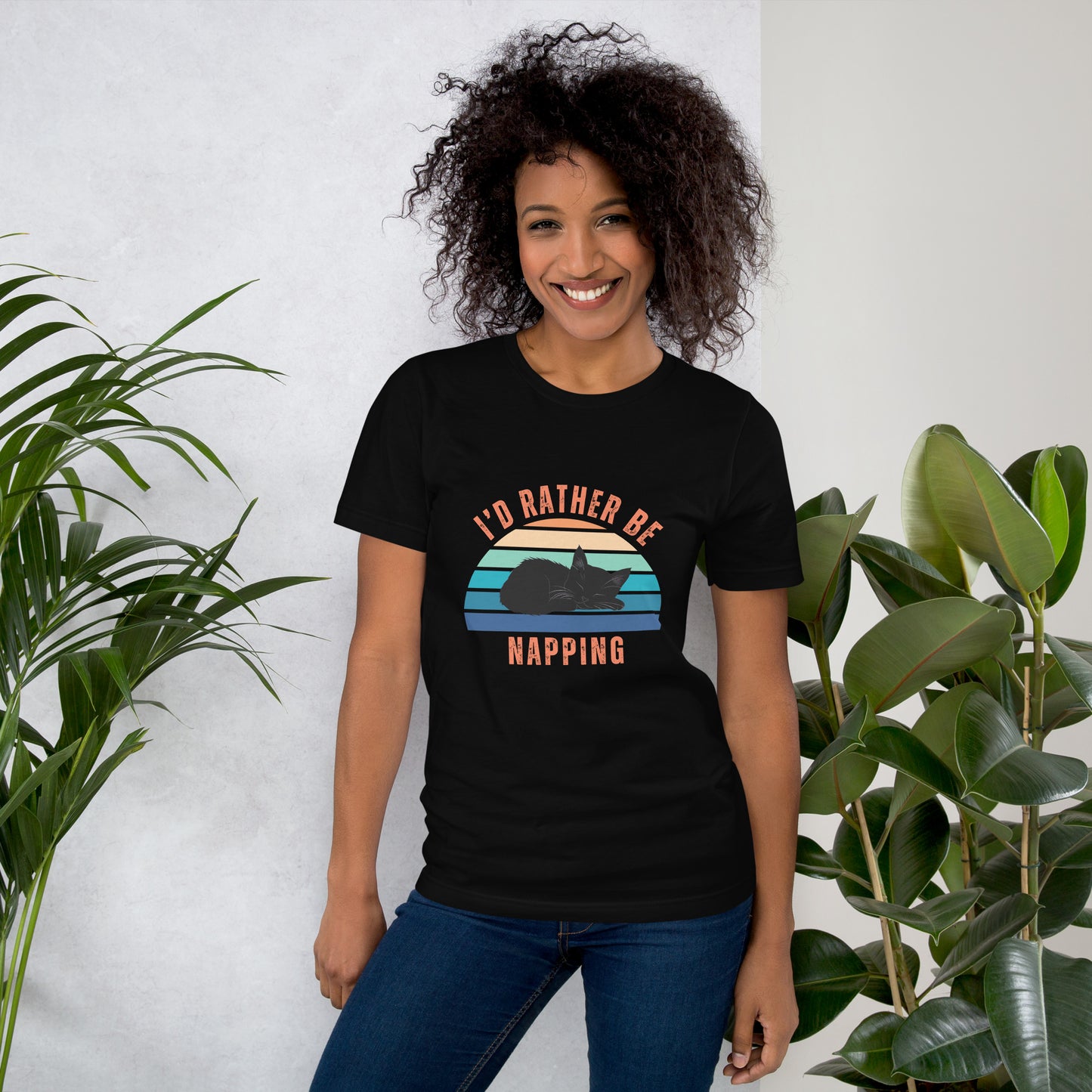 Smiling woman wearing a black unisex t-shirt with a retro-style “I’d Rather Be Napping” design featuring a black cat and sunset stripes, standing near green indoor plants.
