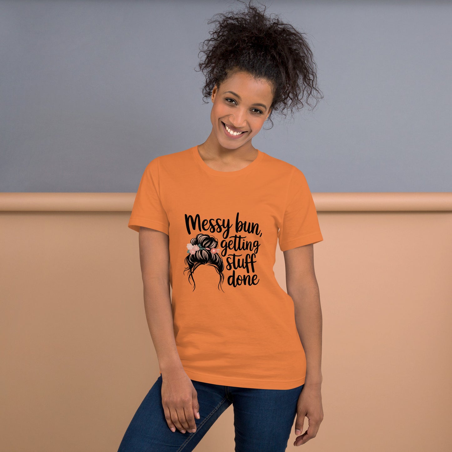 Smiling woman wearing a burnt orange t-shirt with black text that says 'Messy Bun, Getting Stuff Done' and a messy bun illustration