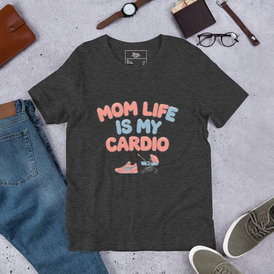 Dark grey unisex t-shirt laid flat on a surface next to jeans and accessories, featuring the phrase 'Mom Life Is My Cardio' in pastel letters with sneakers and pacifier graphic.
