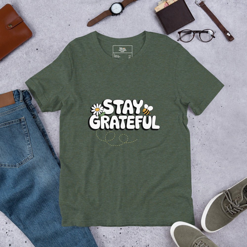 Forest green t-shirt with “Stay Grateful” text design, featuring daisies and a bee trail, styled flat with jeans, sneakers, glasses, a watch, and leather accessories on a gray background.