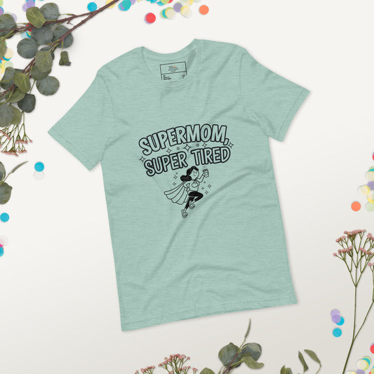 Flat lay photo of a dusty blue t-shirt with a playful "Supermom, Super Tired" comic-style design featuring a flying tired mom holding coffee. Surrounded by eucalyptus leaves and flowers.