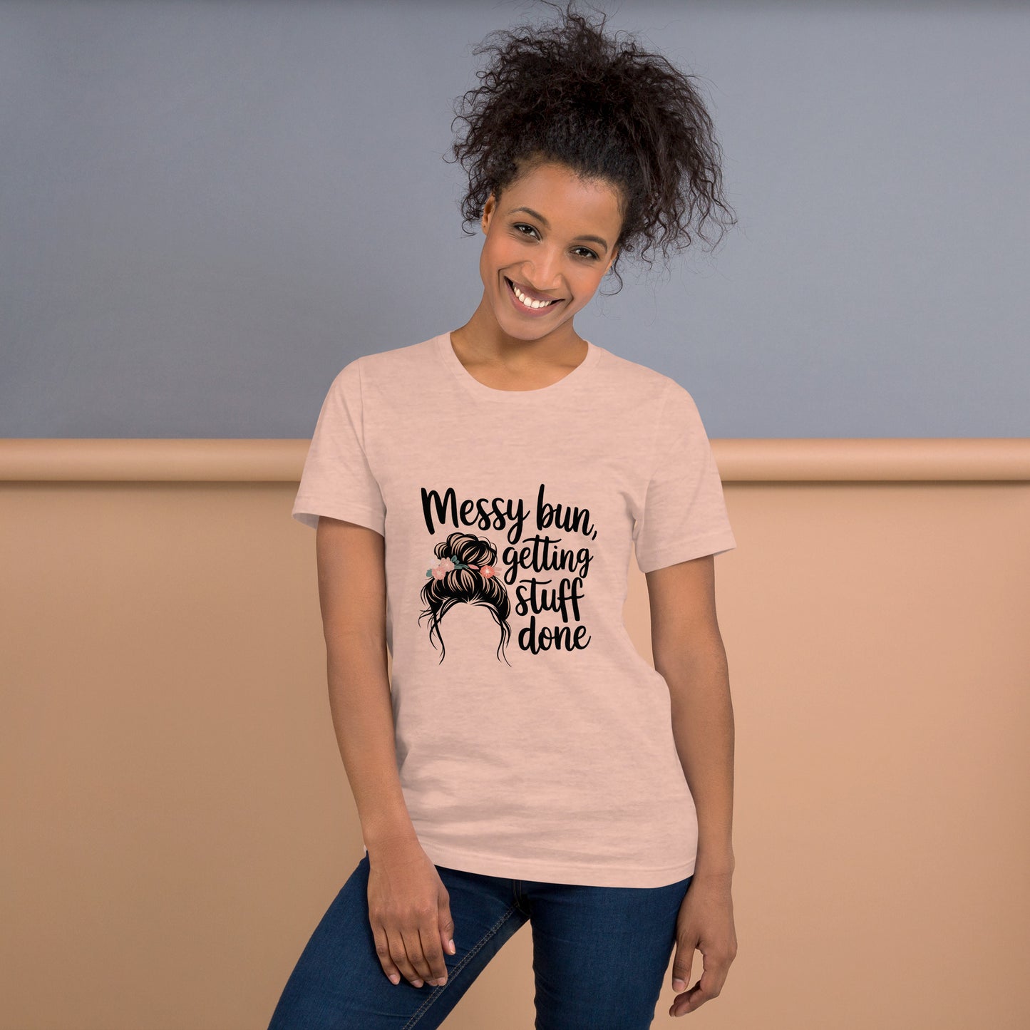 Smiling woman wearing a heather peach t-shirt with black text that says 'Messy Bun, Getting Stuff Done' and a messy bun illustration