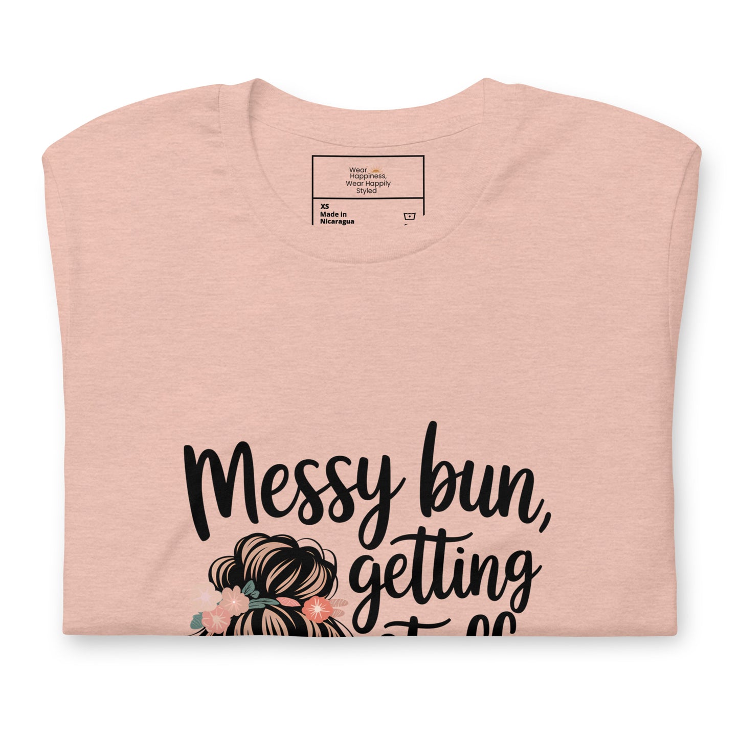 Folded heather peach t-shirt showing black handwritten text 'Messy bun, getting stuff done' with a messy bun illustration and floral details