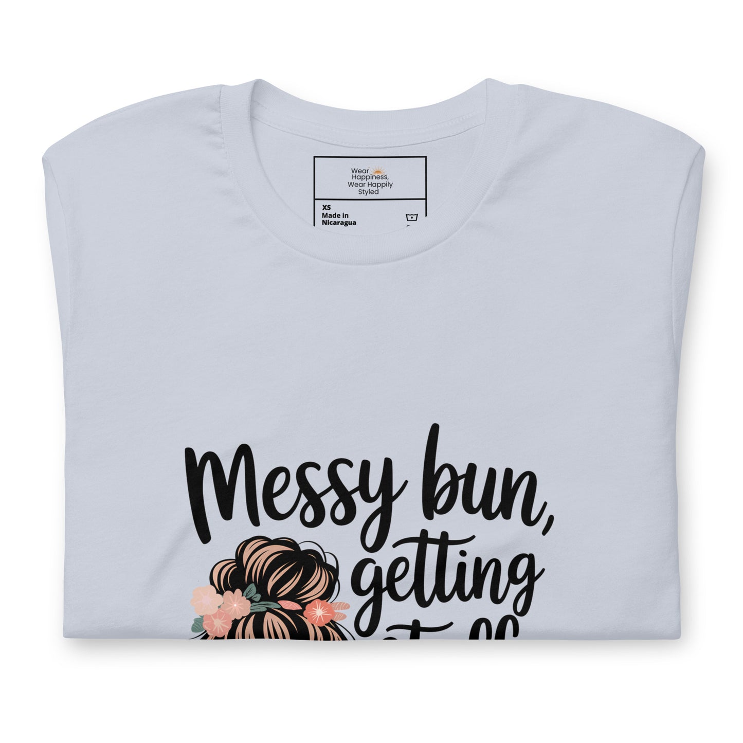 Folded light blue t-shirt showing black handwritten text 'Messy bun, getting stuff done' with a messy bun illustration and floral details
